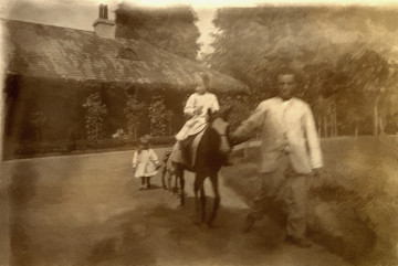 Fotografia przedstawia kilkuletnią dziewczynkę na kucu. Na drugim planie widoczne jest drugie dziecko w towarzystwie psa.