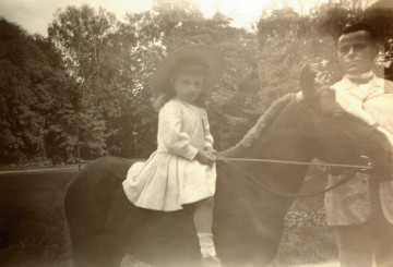 Na fotografii znajduje się 3-4 letni chłopiec w letnim ubraniu i w słomkowym kapeluszu na grzbiecie kuca.