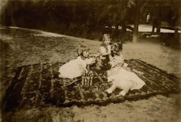 Fotografia przedstawia dzieci siedzące na kocu w przypałacowym ogrodzie a pośrodku nich leży pies.
