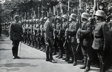 Na zdjęciu znajdują się żołnierze stojący na baczność, prezentujacych broń, w rzędzie; przed nimi stoi dwóch mężczyzn zwróconych do nich twarzą. 