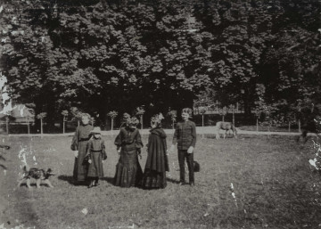 Na zdjęciu znajduje się piec osób, w tym dziecko; z boku stoi pies; w pewnej odległości widać pasącego się konia a w tle drzewa