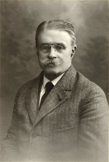 Zdjęcie przedstawia portret mężczyzny w okularach, z wąsami; włosy przeczesane na lewą jego stronę; ubrany w garnitur, biała koszulę i krawat. 