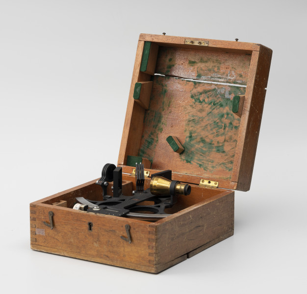 Sekstant Kelvin & Hughes - ujęcie z boku; Sekstant z lunetką w drewnianym pudełku. Sekstant zbudowany z czarnej, metalowej, odlewanej ramy.