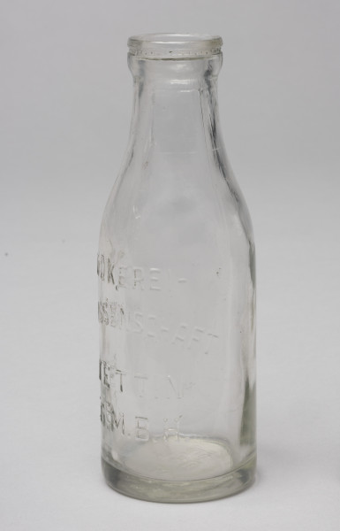butelka po mleku - Ujęcie z boku; Butelka po mleku o pojemności 0,5 litra z przezroczystego szkła z wypukłym napisem. Szyjka butelki krótka, kołnierz wlewu gruby.
