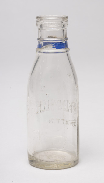 butelka - Ujęcie z tyłu; Butelka po mleku o pojemności 0,5 litra z przezroczystego szkła z wypukłym napisem po drugiej stronie w centralnej części korpusu i niebieskim paskiem w dolnej części szyjki pomiędzy wypukłymi otokami. Szyjka krótka, kołnierz wylewu gruby.