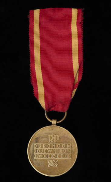 Medal za Warszawę 1939-1945 - ujęcie z tyłu; Okrągły krążek, na którego rewersie umieszczony jest napis w czterech wierszach oddzielonych poziomymi liniami: RP / OBROŃCOM / BOJOWNIKOM / OSWOBODZICIELOM, a pod nim dwa liście dębu. Medal posiada czerwoną wstążkę z żółtymi paskami po bokach.