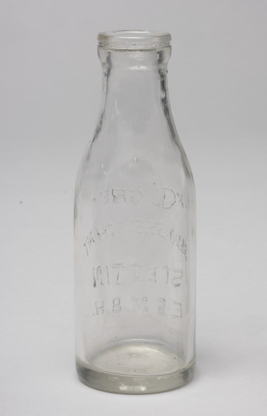 butelka po mleku - Ujęcie z tyłu; Butelka po mleku o pojemności 0,5 litra z przezroczystego szkła z wypukłym napisem po przeciwnej stronie: MOLKEREI GENOSSENSCHAFT STETTIN E.G.M.B.H. Szyjka butelki krótka, kołnierz wlewu gruby.