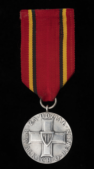 Medal za Udział w Walkach o Berlin - ujęcie z przodu; Oznaką medalu jest wykonany z metalu srebrzonego i oksydowanego krążek, na którego awersie umieszczony jest centralnie wizerunek orderu Krzyża Grunwaldu otoczony na krawędzi koła napisem: ZA UDZIAŁ W WALKACH O BERLIN. Medal zawieszony jest na czerwonej wstążce, z dwoma czarno-żółtymi paskami po bokach. Zawieszenie z przylutowanego uszka z kółeczkiem.