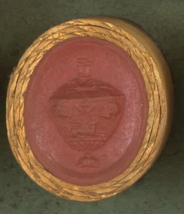 czerwona okrągła gemma w grubym złotym obramowaniu; waza grecka z dekoracją mitologiczną; ma wąską szyjkę, szeroki brzusiec i poprzeczny uchwyt  pod uchwytem leżący sfinks, po obu stronach uchwytu maski, powyżej pegaz.