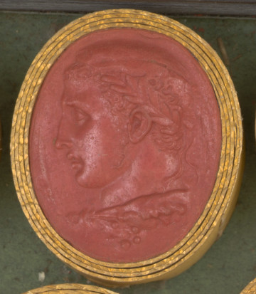 czerwona owalna gemma w grubym złotym obramowaniu, lewy profil młodego mężczyzny z krótkimi kręconymi włosami i wieńcem laurowym na głowie