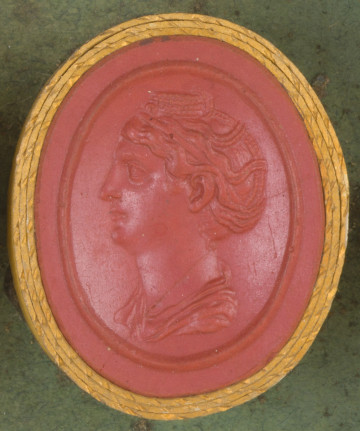czerwona owalna gemma w grubym złotym obramowaniu; lewy profil kobiety o włosach zaplecionych w warkocze i wpiętych na czubku głowy w okrągły kok; poniżej widoczny fragment szaty.