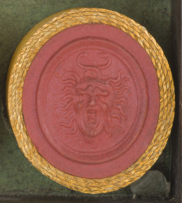 czerwona owalna gemma w grubym złotym obramowaniu; Meduza ma groźny wyraz twarzy, otwarte usta i zmarszczone brwi, jej twarz otaczają grube 