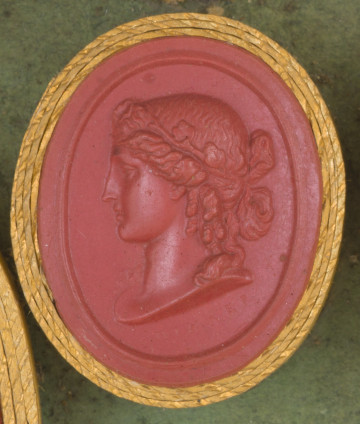 czerwona owalna gemma w grubym złotym obramowaniu; widoczny lewy profil, postać ma włosy upięte w kok i opaskę na głowie, jeden lok spływa na ramię