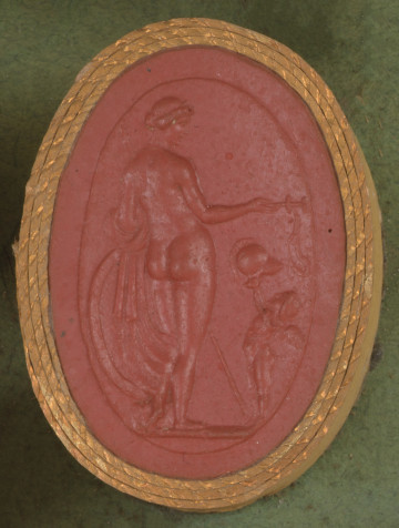 czerwona owalna gemma w grubym złotym obramowaniu; widoczne dwie postaci: po lewej naga kobieta (Wenus) stojąca tyłem do widza, w lewym ręku trzyma płaszcz, w prawym podłużny przedmiot z pętlą (broń Marsa); po prawej stoi skrzydlate putto wręczające kobiecie hełm