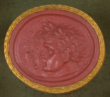 czerwona owalna gemma w grubym złotym obramowaniu; trzy nachodzące na siebie lewe profile głów. Na pierwszym planie widoczny mężczyzna z krótkimi włosami, z wieńcem laurowym na głowie, za nim mężczyzna z krótkimi włosami (bez widocznego wieńca), trzecia postać niewyraźna - z krótkimi włosami i opaską na głowie