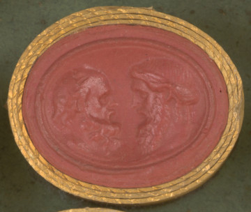 czerwona owalna gemma w grubym złotym obramowaniu; głowy dwóch dojrzałych mężczyzn zwrócone do siebie, widoczne z profilu; mężczyzna po lewej jest łysy ima długą kręconą brodę, mężczyzna po prawej ma długie, częściowo upięte włosy oraz długą brodę i wąsy