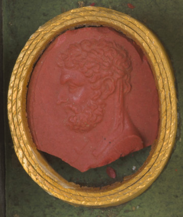 czerwona owalna gemma w grubym złotym obramowaniu; lewy profil mężczyzny w średnim wieku, z krótkimi kręconymi włosami i opaską na głowie, brodą i wąsami; gemma jest uszkodzona: dolna część została odłamana