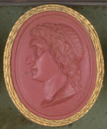 czerwona owalna gemma w grubym złotym obramowaniu, llewy profil głowy młodego mężczyzny z długimi pofalowanymi włosami związanymi opaską 