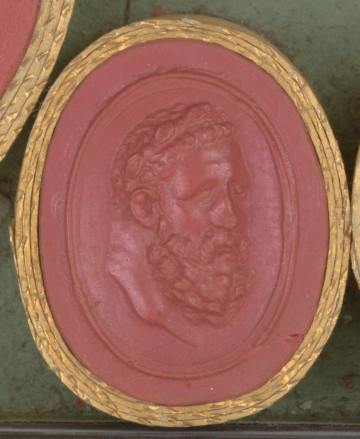 czerwona owalna gemma w grubym złotym obramowaniu, głowa mężczyzny w dojrzałym wieku widoczna z prawego półprofilu; mężczyzna ma krótkie kręcone włosy, wąsy i brodę oraz wieniec laurowy na głowie