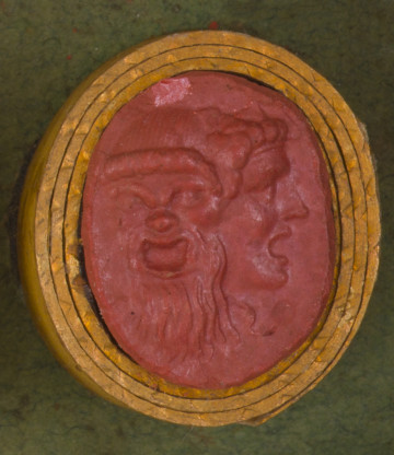 czerwona owalna gemma w grubym złotym obramowaniu; dwie maski groteskowe połączone razem, maska po lewej przedstawia brodatego starca, maska po prawej młodzieńca