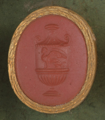 czerwona owalna gemma w grubym złotym obramowaniu; waza z siedzącym gryfem na brzuścu, dekoroacyjnie żłobionym dnem i stopką, na nim dekoracyjna pokrywa w kształcie wklęsłego stożka z osadzoną na szczycie 
