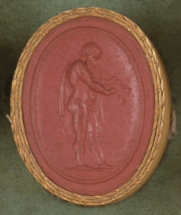 czerwona owalna gemma w grubym złotym obramowaniu; nagi starszy mężczyzna z peleryną zarzuconą na lewe ramię gra na podwójnym flecie (aulosie); postać widoczna jest z prawego boku, ma średniej długości włosy, wysokie czoło i brodę 