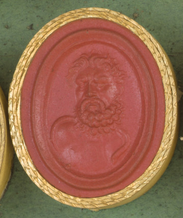 czerwona owalna gemma w grubym złotym obramowaniu; popiersie mężczyzny w średnim wieku, widziane od frontu, mężczyzna ma rozwiane długie kręcone włosy, kręcona brode i wąsy, a na nagich barkach przewieszonego węża.