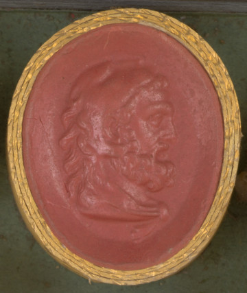 czerwona owalna gemma w grubym złotym obramowaniu; głowa mężczyzny w średnim wieku widoczna z prawego profilu; mężczyzna ma długie kręcone włosy, brodę i wąsy oraz lwią skórę założoną na głowę