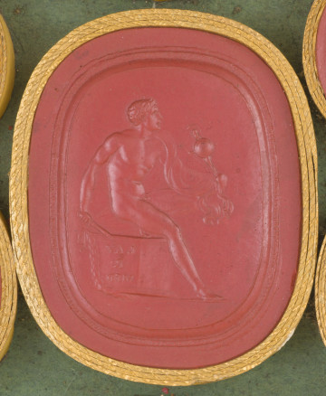 czerwona owalna gemma w grubym złotym obramowaniu; nagi mężczyzna siedzący na postumencie, na którym widoczna jest inskrypcja MED-R-LAV; mężczyzna ma płaszcz zawieszony na lewym ramieniu; w lewej ręce trzyma figurkę Palladium (Atena Pallas), w drugiej ręce sztylet