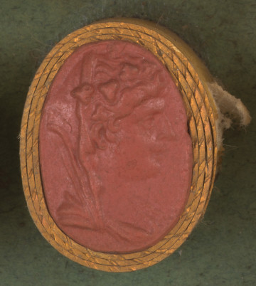 owalna pionowa, czerwona gemma w grubym złotym obramowaniu. Prawy profil młodej kobiety, ma kręcone włosy i kaptur narzucony na tył głowy, na głowie wieniec z winorośli; niżej widoczny fragment szaty