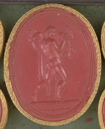 czerwona owalna gemma w grubym złotym obramowaniu, stojący nagi mężczyzna dźwigający zabitego byka na lewym ramieniu, mężczyzna ma lwią skórę przerzuconą przez ramię, scena widoczna z boku.