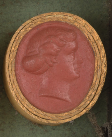 czerwona owalna gemma w grubym złotym obramowaniu; prawy profil kobiety o długich włosach spiętych w kok, z tyłu szyi sygnatura 