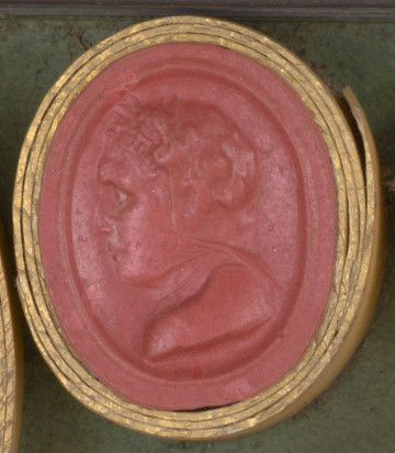 czerwona owalna gemma w grubym złotym obramowaniu, lewy profil popiersia mężczyzny, jego tylną część głowy, nos i usta przykrywa cienka woalka