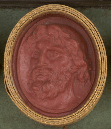 czerwona owalna gemma w grubym złotym obramowaniu; głowa mężczyzny o bujnych kręconych włosach, brodzie i wąsach