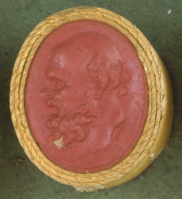 czerwona owalna gemma w grubym złotym obramowaniu; lewy profil starszego mężczyzny z długą, kręconą brodą, wąsami i znaczną łysiną, widoczne krótkie kręcone włosy z tyłu głowy