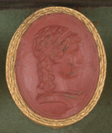 czerwona owalna gemma w grubym złotym obramowaniu; prawy profil chłopca z długimi kręconymi włosami i opaską na głowie, niżej widoczny fragment szaty
