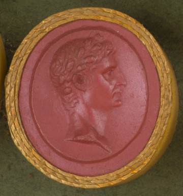 czerwona okrągła gemma w grubym złotym obramowaniu; mężczyzna w średnim wieku z krótkimi włosami i wieńcem laurowym na głowie, widziany z prawego profilu