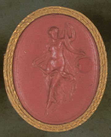 czerwona owalna gemma w grubym złotym obramowaniu; naga młoda kobieta trzyma w rękach zwiewną tunikę, którą częściowo okrywa swoje ciało; ma włosy spięte w kok, a na szyi i na ręku sznur z pereł, wydaje się robić krok w stronę widza