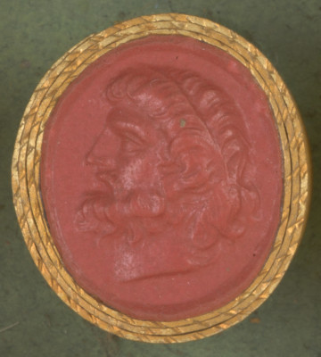 czerwona owalna gemma w grubym złotym obramowaniu, mężczyzna w średnim wieku widziany z lewego profilu, ma długie włosy z nałożoną cienką opaską, wąsy i brodę oraz srogi wyraz twarzy
