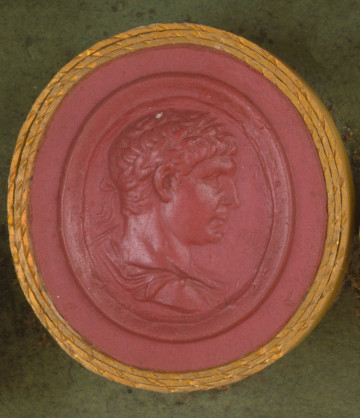 czerwona okrągła gemma w grubym złotym obramowaniu; prawy profil mężczyzny w średnim wieku, ma krótkie włosy i wieniec laurowy na głowie, w dolnej części widoczny fragment szaty 