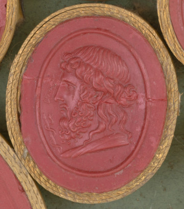 czerwona owalna gemma w grubym złotym obramowaniu, widoczny lewy profil mężczyzny z długimi włosami częściowo spiętymi wokół głowy i z długą kręconą brodą i wąsami; obok widoczna laska w wijącym się wokół niej węzem.