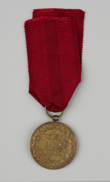 Medal Pamiątkowy Za Wojnę 1918-1921 - rewers; Okrągły medal z oczkiem do zawieszania na górze. W polu rewersu znajduje się dewiza w trzech wierszach: POLSKA SWEMU OBROŃCY, otoczona wieńcem z liści dębowych. Medal zawieszony na zastępczej wstążce w kolorze czerwonym.