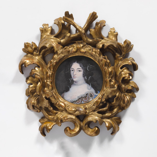 Fotografia kolorowa. Owalna rama w kolorze złotym. W otoku ozdobiona motywem liści, które rozchodzą się tworząc kolejny krąg z tym samym motywem. Pośrodku portret młodej kobiety. Twarz owalna, blada. Duże ciemne oczy, z łagodnie zarysowanymi łukami brwiowymi. Usta pełne. Włosy ciemne, z przedziałkiem pośrodku. Na odsłonięte czoło z dwóch stron spadają spiralne loki. We włosy wplecione ozdoby ze sznurów białych pereł. Taki sam krótki sznur pereł obejmuje szyję kobiety. Głęboki dekolt wykończony koronką, spod której widoczne są naszyte na suknię ozdoby z pereł i fragment materiału w kolorze niebieskim i złotym.