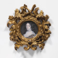 Fotografia kolorowa. Owalna rama w kolorze złotym. W otoku ozdobiona motywem liści, które rozchodzą się tworząc kolejny krąg z tym samym motywem. Pośrodku portret młodej kobiety. Twarz owalna, blada. Duże ciemne oczy, z łagodnie zarysowanymi łukami brwiowymi. Usta pełne. Włosy ciemne, z przedziałkiem pośrodku. Na odsłonięte czoło z dwóch stron spadają spiralne loki. We włosy wplecione ozdoby ze sznurów białych pereł. Taki sam krótki sznur pereł obejmuje szyję kobiety. Głęboki dekolt wykończony koronką, spod której widoczne są naszyte na suknię ozdoby z pereł i fragment materiału w kolorze niebieskim i złotym.