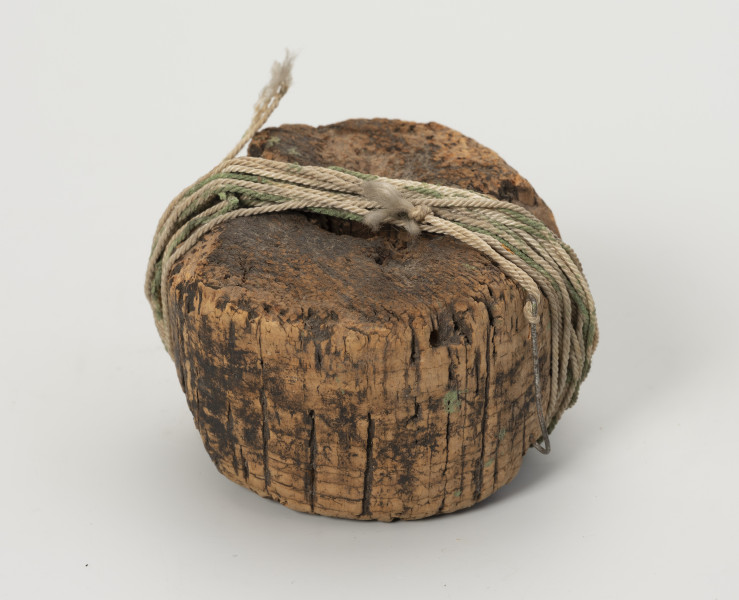 węda pływająca - Ujęcie z przodu; Węda pływająca składająca się z okrągłego pływaka z otworem w środku, wykonanego z korka, na powierzchni którego widać liczne obłupienia. Do pływaka przywiązana jest bawełniana nić, na końcu której umieszczony jest metalowy haczyk.