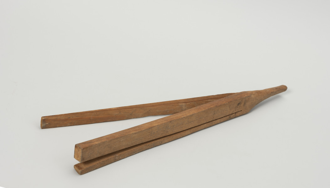 klamra do haczyków - Ujęcie z przodu; Drewniana klamra ma kształt podłużnej listewki o czworobocznym przekroju. jeden z końców rozszczepiono do 3/4 długości. Drugi zaostrzono na końcu nadając mu stożkowaty kształt pasujący do otworu w ramieniu stojaka, w które wkładano sznurki z zawieszonymi na nich zestawami haczyków do suszenia.