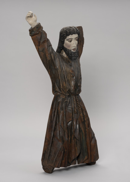 tłok pieczętny - Ujęcie ze skosu z lewej; Męska figura zakomponowana frontalnie w pozycji stojącej na kształt litery X, z uniesionymi rękami i rozstawionymi nogami. Przedstawia młodego mężczyznę ubranego w długą obszerną tunikę zapinaną na guziki i spiętą w pasie sznurem. Światłocieniowe fałdowania szaty zostały oddane naturalistycznie, zgodnie z układem ciała. Tunika powyżej paska rozchyla się. Podłużna twarz o wyrazistych kościach policzkowych oraz dużych ustach i oczach. Fryzura potraktowana dekoracyjnie. Tworzą ją ciemne, wyraziste spirale półdługich włosów, krótka grzywka i średniej długości broda. Figura przyścienna z tyłu płaska. Stopy i lewa dłoń nie zachowały się.