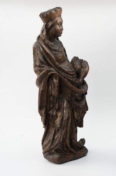 Piękna Madonna z Żarnowa - Ujęcie ze skosu z lewej; figura ołtarzowa; Kobieca figura stojąca na czworobocznej podstawie w obszernych szatach i koronie, trzymająca przy boku dzieciątko. Sylwetka kobieca zakomponowana w wyraźnym kontrapoście, odchylająca tors do tyłu i w lewo, z twarzą zwróconą w trzech czwartych w prawo ku dzieciątku trzymanym na lewym biodrze. Maria ukazana w wieku młodzieńczym nosi długą suknię i płaszcz, którego poła przerzucona przodem układa się w miękką kolistą, wydatną fałdę. Brzegi płaszcza opadają poniżej uniesionych do przodu dłoni spiętrzonymi dekoracyjnie fałdowaniami. Na głowie nałożona wysoka korona oraz chusta zsuwająca się do tyłu i opadająca miękkimi łukami wokół szyi. Twarz owalna z wysokim czołem i wydatnymi policzkami, o łagodnym wyrazie. Dzieciątko o pulchnych nóżkach siedzi przy biodrze, podtrzymywane lewą dłonią matki. Brakuje prawej dłoni Marii i figury dzieciątka powyżej bioder. Na powierzchni rzeźby widoczne pozostałości polichromii.