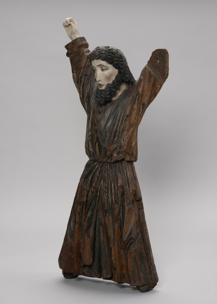 tłok pieczętny - Ujęcie ze skosu z prawej; Męska figura zakomponowana frontalnie w pozycji stojącej na kształt litery X, z uniesionymi rękami i rozstawionymi nogami. Przedstawia młodego mężczyznę ubranego w długą obszerną tunikę zapinaną na guziki i spiętą w pasie sznurem. Światłocieniowe fałdowania szaty zostały oddane naturalistycznie, zgodnie z układem ciała. Tunika powyżej paska rozchyla się. Podłużna twarz o wyrazistych kościach policzkowych oraz dużych ustach i oczach. Fryzura potraktowana dekoracyjnie. Tworzą ją ciemne, wyraziste spirale półdługich włosów, krótka grzywka i średniej długości broda. Figura przyścienna z tyłu płaska. Stopy i lewa dłoń nie zachowały się.