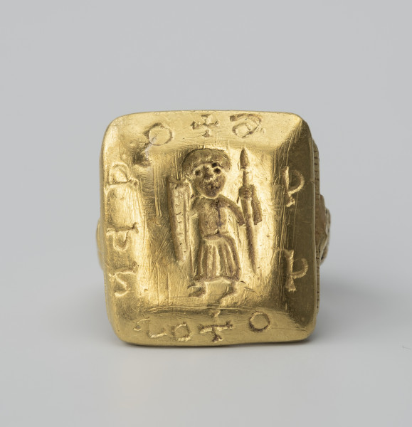 pierścień z Pęzina - Ujęcie z przodu; Pierścień zbudowany z obrączki i dużej prostokątnej tarczki. Tarczka prostokątna, zbliżona do kwadratu, o fasetowanych bokach. W środkowej części tarczki, we wklęsłym reliefie, postać stojącego mężczyzny w długiej szacie, z włócznią i podłużną, trójkątną tarczą w dłoniach. Na sfazowanych brzegach tarczki nierozpoznana inskrypcja, składająca się z 10 znaków rozdzielonych dwoma znakami krzyża greckiego.